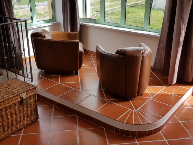 2 fauteuils Club BATEAU-CIGARE marron Rochembeau face à une baie vitrée donnant sur jardin et à côté d'une malle en osier