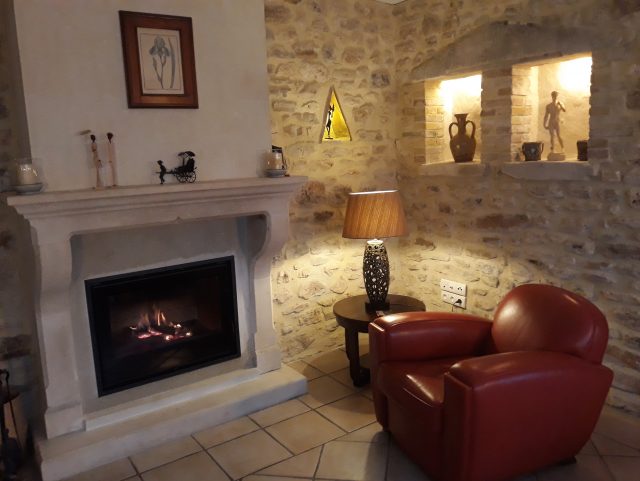 Fauteuil Club cuir rouge Rochembeau à côté d'un feu de cheminée, et mur en pierres claires