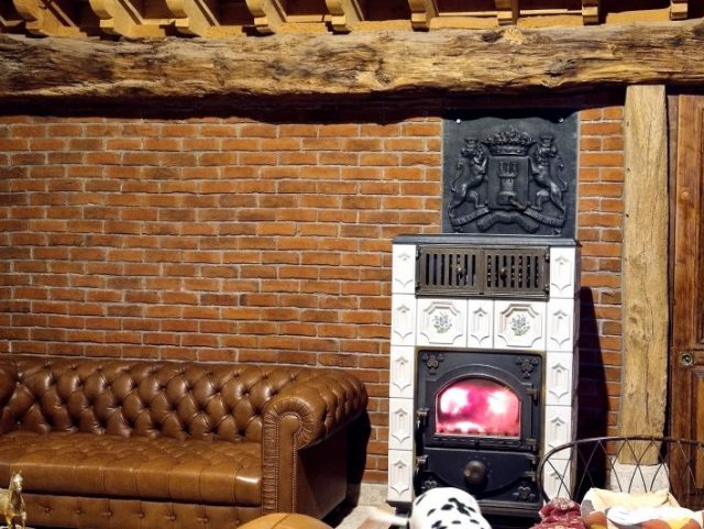 Canapé Chesterfield cuir marron Rochembeau, mur en brique, poêle à bois, grosse poutre et porte en bois 